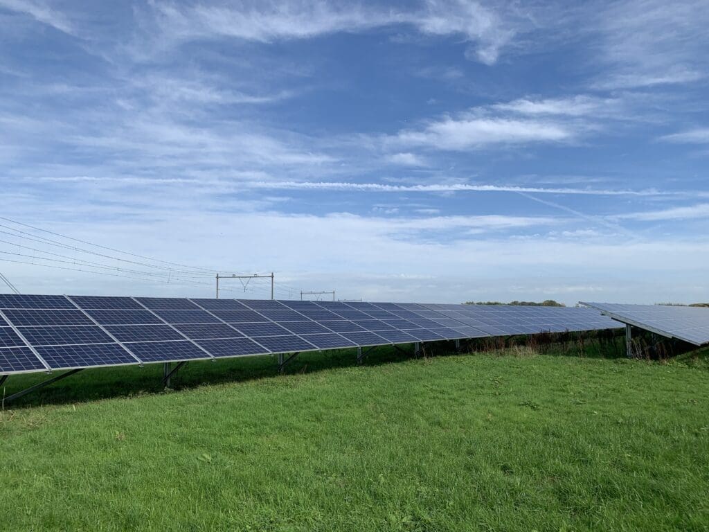 Zonnepark op een graslandlocatie in Nederland met rijen grondgebonden zonnepanelen en hoogspanningsmasten op de achtergrond